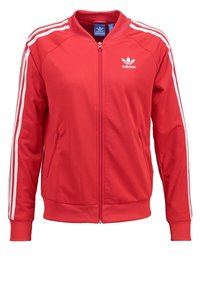 Červená sportovní bunda Adidas s bílými třemi pruhy na rukávech, žebrovanými manžetami, předním zipem a postranními kapsami. Hladký, lehký materiál.