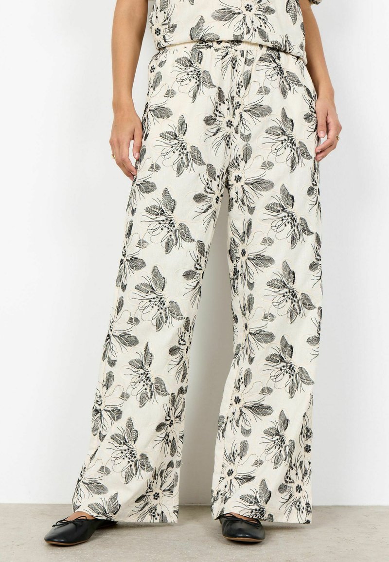 Femme portant un pantalon large crème avec un grand imprimé floral noir et des ballerines noires, debout contre un mur blanc.