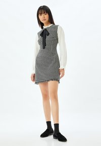 Sort-hvid houndstooth kjole med flæsekrave og sort sløjfedetails, parret med sorte loafers og sokker, frynsesøm og tætsiddende pasform.