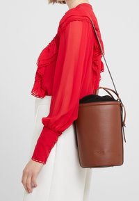 Borsa a secchiello in pelle marrone con rifinitura strutturata, fodera nera e tracolla lunga, abbinata a una blusa rossa con rouches e dettagli sui bottoni.