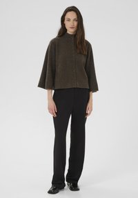 Egy nő barna, gombos gyapjú pulóverben, széles ujjal, fekete egyenes nadrágban és fekete loaferekben áll a fehér háttér előtt.