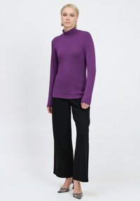 Lila langärmeliger Rollkragenpullover kombiniert mit schwarzen weit geschnittenen Hosen und gemusterten High Heels, vor einem neutralen Hintergrund präsentiert.