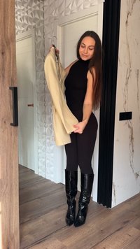 Mujer con vestimenta negra sin mangas y botas negras brillantes sosteniendo una chaqueta beige en una percha dentro de una habitación moderna.