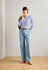 Stripet langermet skjorte i hvit og blå, lyseblå rutete jeans, brun belte, brune hæler, og en brun jakke slengt over den ene armen.