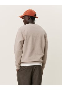 Beige sweatshirt met een ronde hals, textuurstof en geribbelde manchetten, gecombineerd met een roestkleurige pet. Zichtbaar wit t-shirt eronder.