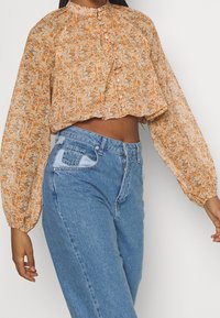 Blusa corta de manga larga con estampado floral ligero en tonos naranjas y crema. Cuenta con un escote volado y un frente de botones. Combinada con jeans azules.
