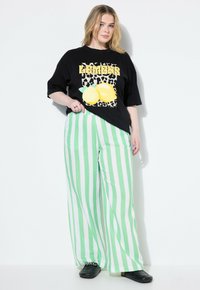 Svart grafisk t-shirt med citrusskrift och leopardbakgrund; parat med vida, vertikala grön- och vitrandiga byxor. Svarta platta skor.