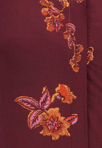 Βουργουνδικό ύφασμα με λουλουδάτο μοτίβο σε πορτοκαλί και ροζ, με υφαντές φύλλα και περίπλοκες λεπτομέρειες κατά μήκος των άκρων.