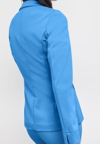 Sininen bleiseri, joka on räätälöity pehmeästä kankaasta ja siinä on vartalonmyötäinen muotoilu, rakenteelliset olkasaumat ja sivuventtiilit liikkuvuuden lisäämiseksi.