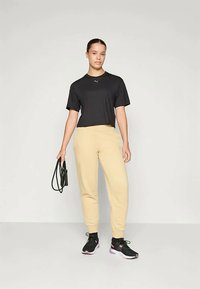 Melnā īsā kreklā, bēžos treniņbiksēs un melnās sporta kurpēs. Modelis tur melnu somu. Vienkārša, brīva piegriezuma un mīksti audumi.