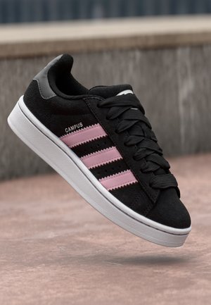 Czarny sneaker Adidas Campus z różowymi paskami i białą podeszwą, zawiązany, umieszczony w powietrzu na tle kamienia i betonu.