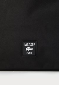 Tessuto nero texturizzato con un logo quadrato nero al centro che riporta la scritta "LACOSTE PARIS" e presenta un'icona di coccodrillo bianca.