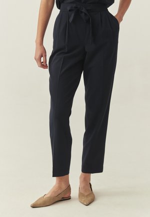 Spodnie w kolorze granatowym, dopasowane, z plisami, o wysokim stanie i paskiem do wiązania. Połączone z beżowymi płaskimi butami z noskiem w szpic, z wycięciem po bokach.