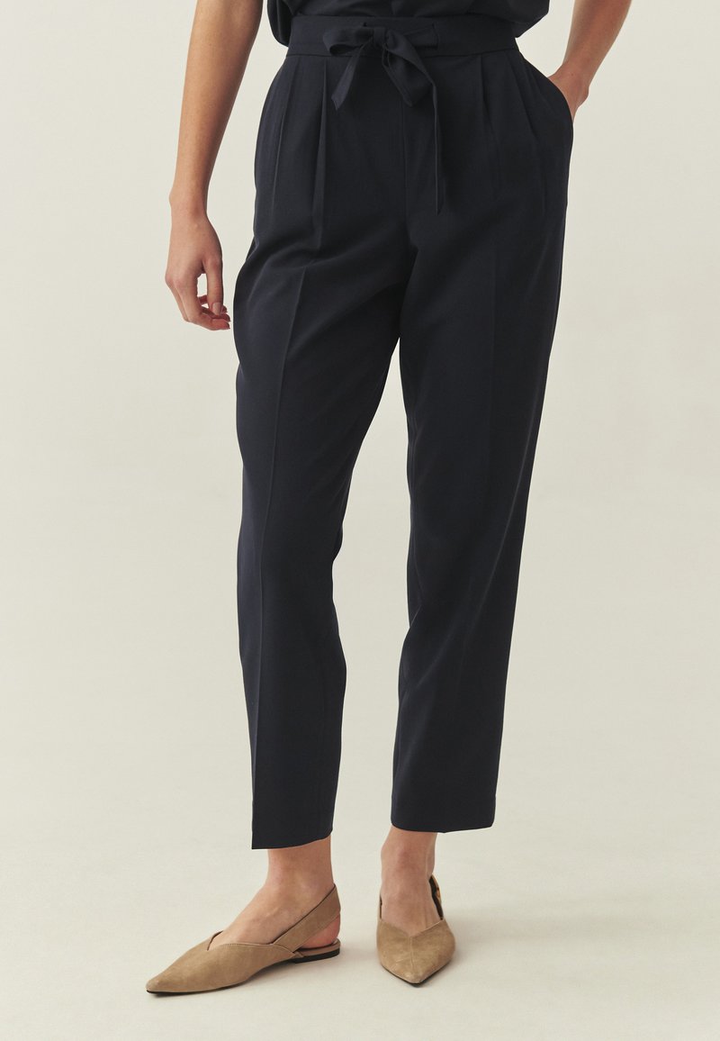 Pantalon marine sur mesure avec plis, taille haute et ceinture à nouer. Associé à des ballerines beiges à bout pointu, avec un design découpé sur le côté.