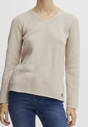 Person trägt einen hellbeigen strukturierten Strickpullover und dunkelblaue Jeans und steht vor einem schlichten hellen Hintergrund.