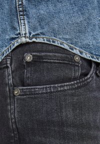 Dunkelblaue Jeans mit einer vorderen Tasche, die mit silberner Hardware versehen ist. Der Stoff hat eine glatte Textur, die durch ein helleres Jeanshemd darüber kontrastiert wird.