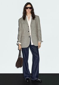 Blazer grigio a quadri oversize, camicia bianca, jeans scuri a zampa di elefante, scarpe lucidate nere, occhiali da sole neri e borsa marrone.
