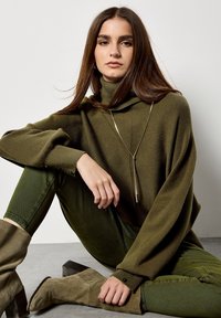 Pull à col roulé kaki à coupe oversize, texture côtelée et fentes sur les côtés, associé à un jean skinny vert foncé et des bottes beiges montant jusqu'aux genoux.