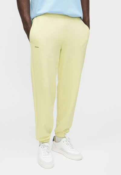 Mężczyzna noszący jasnżółte spodnie dresowe ze ściągaczami, białe trampki i jasnoniebieską koszulę, z rękami w kieszeniach, stoi na jednolitym tle.