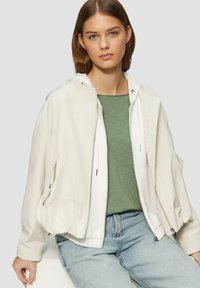 Leichte Bomberjacke aus Cream mit Kapuze, Reißverschluss vorne, zwei Taschen, kombiniert mit grünem Oberteil und hellblauen Jeans. Glatte Stoffstruktur.