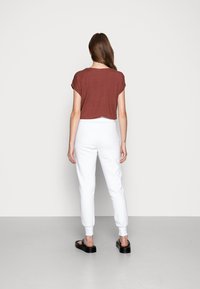 Kvinna som står vänd mot en vit vägg, klädd i en kortärmad brun topp, vita joggerbyxor och svarta plattformssandaler.