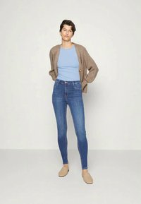 Top ajustado azul claro combinado com um longo cardigan castanho e jeans skinny azuis. A modelo usa sapatos slip-on bege. Design simples e clean.