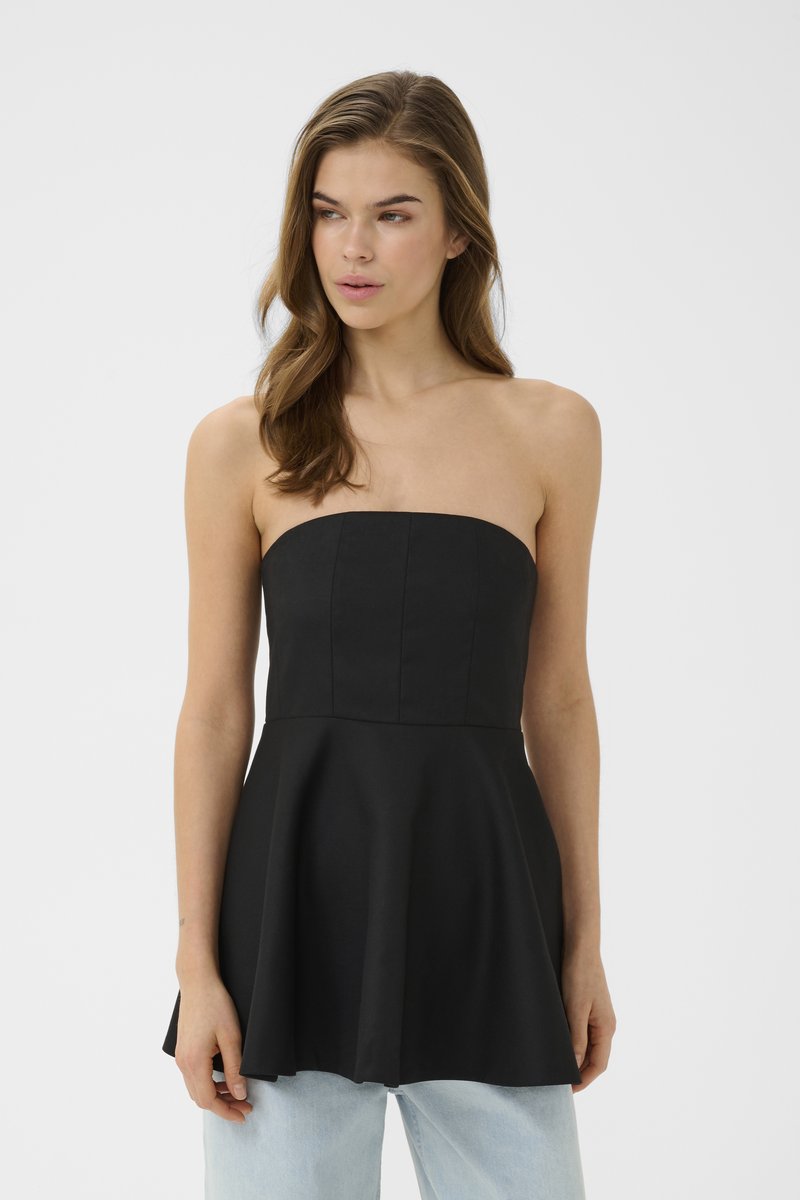 Γυναίκα που φοράει μαύρο τοπ χωρίς τιράντες με φούστα και ανοιχτόχρωμο τζιν, στέκεται μπροστά σε απλό ανοιχτό φόντο, κοιτώντας προς τα αριστερά.