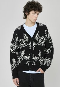Cardigan noir avec un motif floral blanc, en col en V, boutons sur le devant et poignets côtelés. Porté par-dessus un t-shirt bleu clair à coupe décontractée.