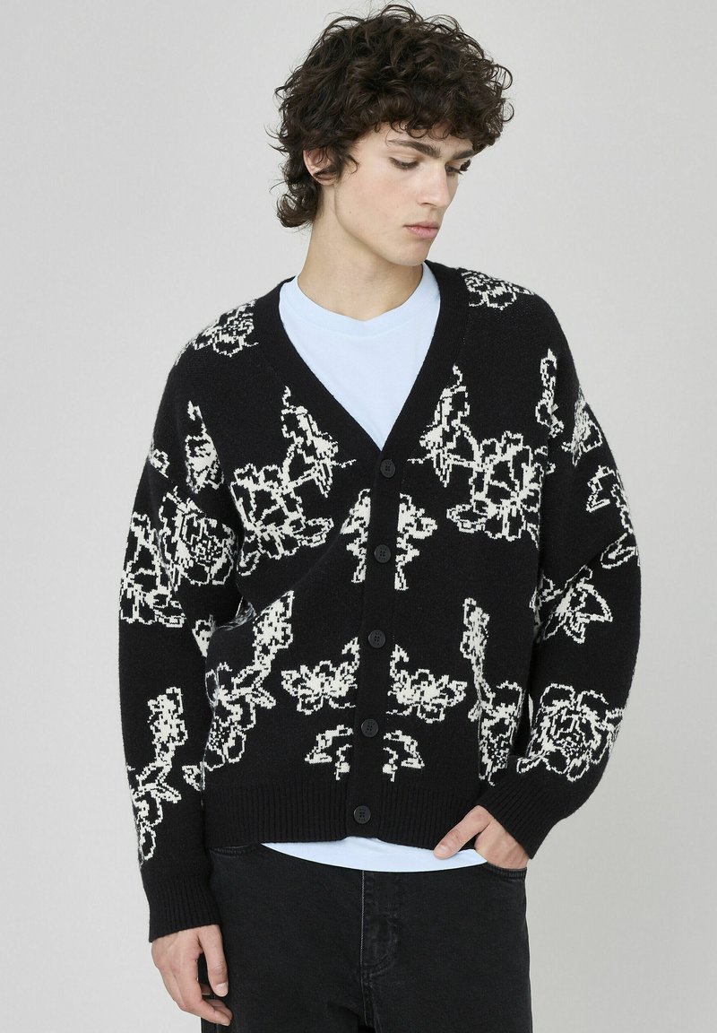 Cardigan noir avec un motif floral blanc, en col en V, boutons sur le devant et poignets côtelés. Porté par-dessus un t-shirt bleu clair à coupe décontractée.