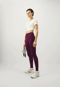 Leggings roxos de cintura alta, um top cropped branco justo e sapatos de atletismo cinzentos. O modelo segura uma corda de pular com uma mão.