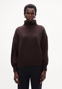 Pullover marrone oversize con collo alto, orlo e polsini a coste, realizzato in tessuto a maglia testurizzato, abbinato a pantaloni scuri a righe sottili.