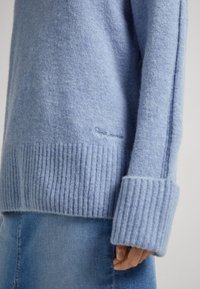 Pull tricoté bleu clair avec poignets et ourlet côtelés, présentant une texture douce et un branding subtil sur le côté.
