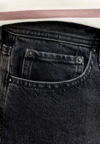 Μαύρα τζιν από ντένιμ με μπροστινή τσέπη που έχει εμφανείς ραφές και μεταλλικά καρφιά. Το τελείωμα ενός ανοιχτόχρωμου πουκαμίσου φαίνεται μερικώς επάνω τους.