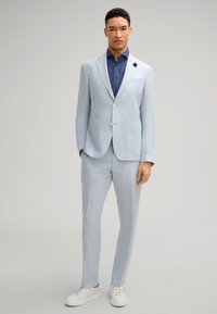 Helles blaues Leinenanzug mit einem einreihigen Blazer und passender Hose, kombiniert mit einem navyblauen Hemd und weißen Sneakers.