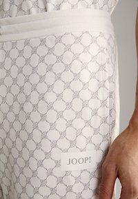 Pantalons de jogging de couleur claire présentant un motif géométrique répétitif, avec un tissu texturé et une étiquette visible portant la mention "JOOP!" sur le côté.