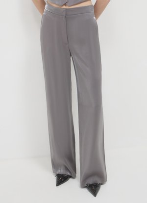 Frau trägt hoch taillierte, weit geschnittene graue Satinhosen, kombiniert mit schwarzen spitzen Schnürschuhen vor weißem Hintergrund.