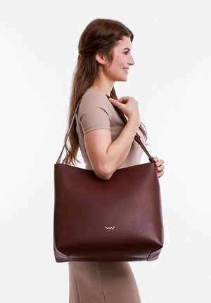 Brown leather tote bag with a smooth texture, a spacious shape, and a logo detail on the front. Worn on the arm by a model.