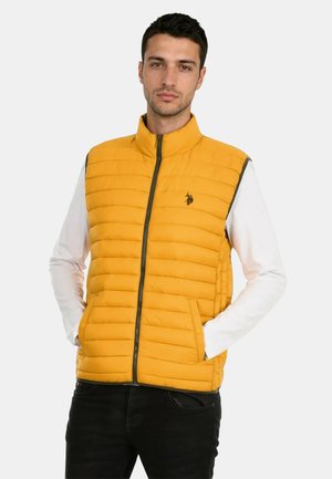 Junger Mann in einer gelben, gesteppte ärmellose Jacke mit Frontreißverschluss, trägt ein weißes Langarmshirt und schwarze Hose, Hände in den Taschen.