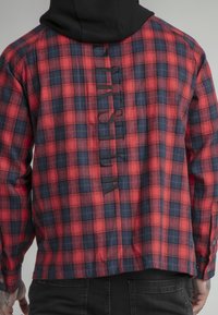 Camicia a quadri rossi e navy con cappuccio nero, presenta un orlo dritto, polsini con bottoni e un dettaglio testuale verticale lungo la schiena.