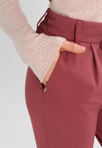 Bordeauxbroeken met een gladde textuur, voorzien van een brede tailleband en zijzakken. Lichte roze, geribbelde trui met een mouw in beeld.