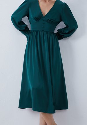 Femme portant une robe midi à manches longues de couleur bleu sarcelle profond, avec des boutons sur la taille cintrée et un décolleté en V.