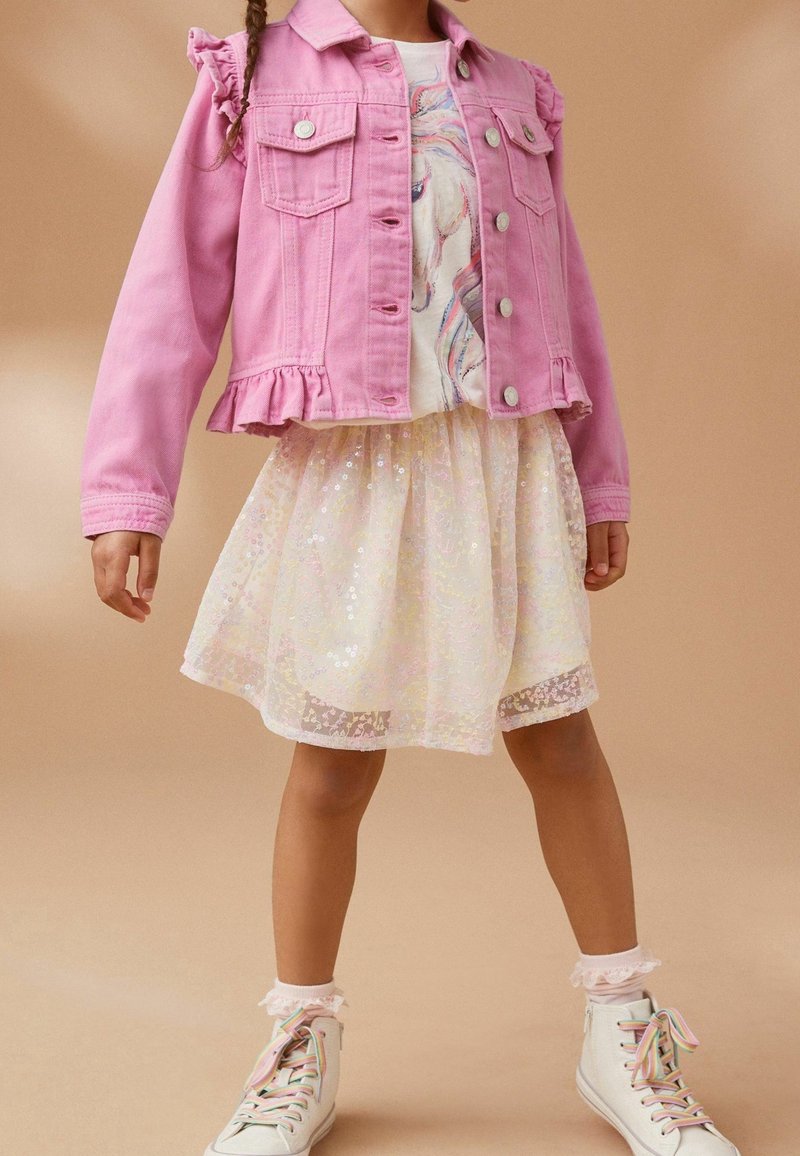 Rosa Jeansjacke mit Rüschenschultern, getragen über einem weißen Oberteil mit einem Einhorn-Design, kombiniert mit einem leichten, glitzernden Rock und weißen Sneakers.