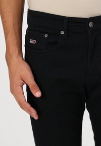 Close-up van de hand van een persoon die naast een paar zwarte spijkerbroeken ligt met een klein rood, wit en blauw logo-opkleefstuk op de voorzak.