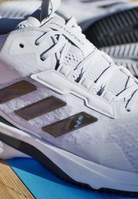 Białe buty sportowe z siateczkową cholewką, trzema ciemnoszarymi paskami i teksturowaną podeszwą. Wyposażone w wyściełany język i sznurowadła zapewniające bezpieczne dopasowanie.