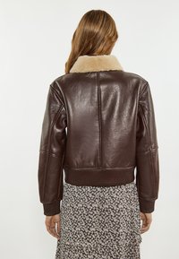 Giacca di pelle marrone con un colletto beige soffice, polsini e orlo a coste, caratterizzata da una texture liscia e lucida e un design aderente.