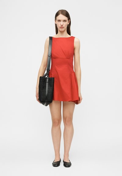 Γυναίκα που φοράει κόκκινο αμάνικο φόρεμα και μαύρα φλατ παπούτσια, κρατώντας μια μεγάλη μαύρη πλεκτή τσάντα ώμου, στέκεται μπροστά σε ένα ανοιχτό γκρι φόντο.