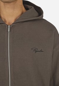 Haut à capuche zippé marron à côtes avec une grande capuche, fermeture éclair argentée et un logo noir discret sur la poitrine gauche. Texture douce et coupe décontractée.