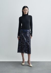 Black long-sleeve top paired with a knee-length, sheer navy floral skirt. Model wears beige high-heeled shoes. Smooth textures and layered design.