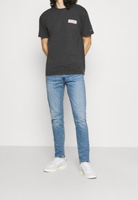 Svart bomullströja med färggrafik av "JEANS" på vänster bröst, kombinerad med ljusblå denimjeans och vita sneakers.