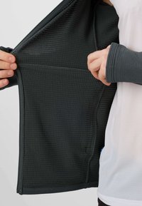 Oseba drži odprto črno teksturirano jakno, prikazuje notranji žep in luknjo za palec na rokavu čez belo majico.